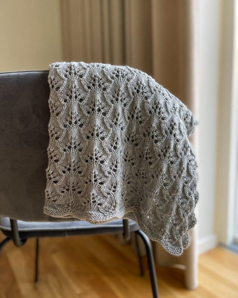 Et gråt Blueberry Baby Blanket i Bøllefrø fv. 1 Lys Grå, hænger over en grå stol i en stue.